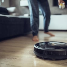 A robot vacuum on a wooden floor. A person's legs are seen running behind it.