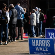 Harris and Trump yard signs are staked next to a line of voters entering a polling place.  