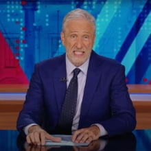 A man in a suit sits behind a talk show desk, grimacing.