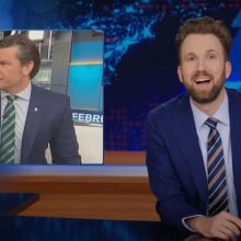 A man sits behind a talk show desk laughing with shock. In the top-left is an image of a man in a suit on TV.