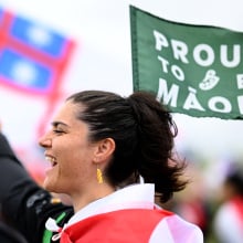 A person cheers and holds up a green flag that reads "Proud to be Maori."