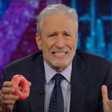 A man in a suit sits behind a talk show desk holding a donut.