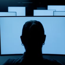 The back of a woman's head, as she stares at several glowing blue screens.