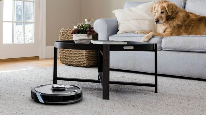 a shark matrix plus robot vacuum cleans the carpet in a living room that also has a table and a dog laying on a couch