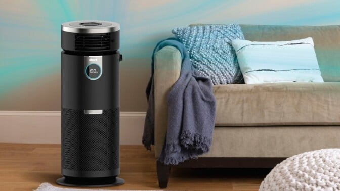 a shark air purifier sits to the left hand side of a beige sofa that has a blue throw blanket over the left arm.