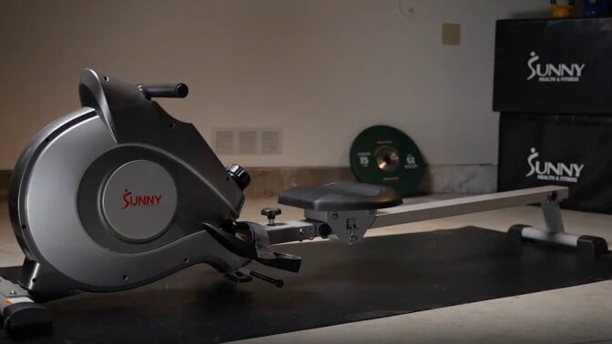 a sunny health rowing machine sits on a black mat on the floor with two large black boxes in the background that say sunny health