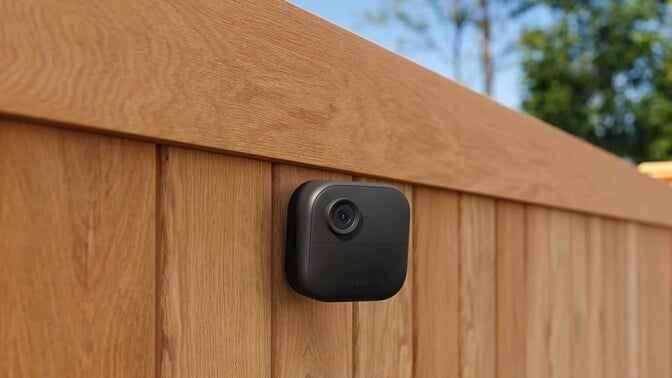 a blink outdoor 4 camera is mounted onto a wooden fence. Behind the fence is blue sky and green trees on a sunny day