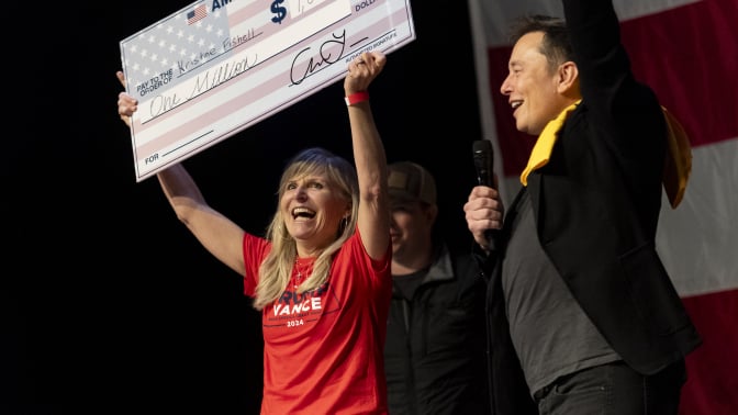 Elon Musk stands on stage cheering for a woman holding up a giant check written out for $1 million.