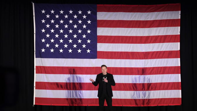 Elon Musk stands on stage in front of a giant American flag.