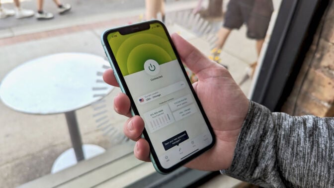 a close-up of a man holding an iphone displaying the expressvpn app in front of a window