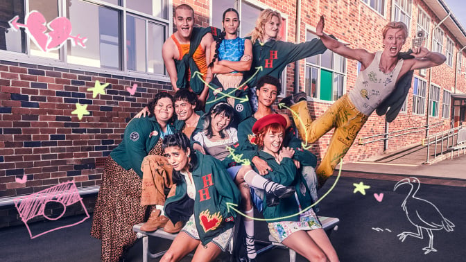 The cast of Netflix's "Heartbreak High" sititng on an aluminium picnic table, wearing Hartley High's green leaver jackets.