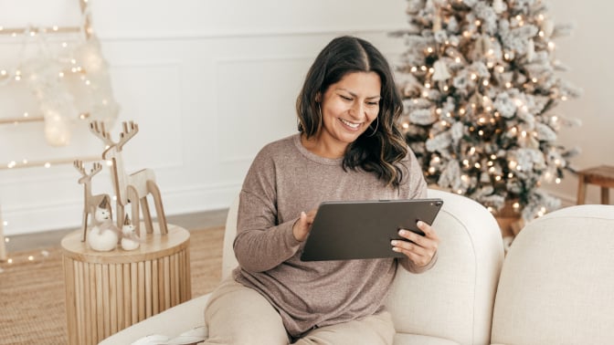 a person sits on a white couch while looking at a tablet and smiling. In the background, a lit up holiday tree