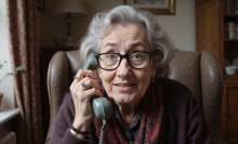 An elderly woman looks into the camera holding a landline telephone.